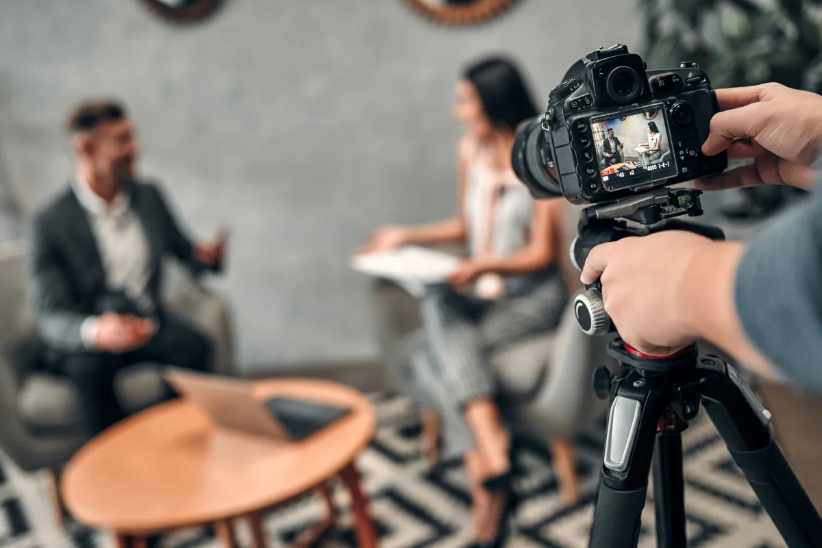 Im Vordergrund ein Stativ mit Kamera, im Hintergrund findet ein Interview statt, unscharf zu sehen sind eine weibliche und eine männliche Person in einer Interviewsituation.